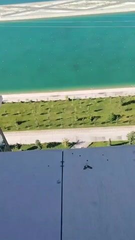 The video shows an aerial view of a large, artificial body of turquoise water, possibly a rowing or swimming channel, with white lines marking lanes. On one side of the water is a manicured green lawn with rows of small trees. Beyond the lawn is a paved path and then a building with a beige facade. Across the water, there are two large, modern hotel-like buildings, one with a blue water slide structure adjacent to it. The landscape in the background includes green fields, some buildings, and mountains in the distance. The sky is clear and blue. The video appears to be shot from a high vantage point, possibly a drone or a tall building.