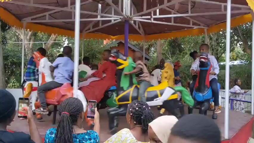 The video captures a group of people, primarily children, enjoying a ride on a merry-go-round. The merry-go-round has a canopy supported by a white frame and topped with a yellow awning. It features a variety of colorful, stationary horses for riders to sit on. The children are sitting on the horses, facing forward and appear to be enjoying the ride.  Several people are standing around the merry-go-round, observing and recording with their phones. The setting appears to be an outdoor park or recreational area. In the background, there are trees, a small building, and a fence. The atmosphere seems lively and cheerful.
