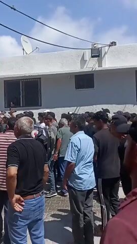 A group of men are gathered in front of a white building. A tall, damaged building with many holes in its exterior is visible in the background. Some of the men are looking towards the damaged building. It appears to be daytime and the sky is clear and blue.