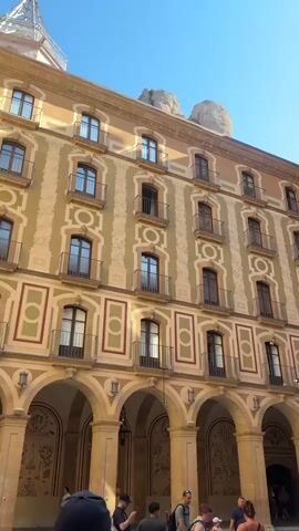 The video shows a courtyard with people and buildings. In the foreground, several people are visible from the chest up, some wearing hats. The background features a multi-story building with arched walkways on the ground floor and balconies on the upper floors. The facade is decorated with a pattern of cream and light green alternating colors, with red and cream trim around the windows. To the right of this building, a large, ornate brick structure with elaborate carvings and a circular window is visible, likely a church or cathedral. The sky is clear and blue. The overall impression is of an historic European setting, possibly a monastery or religious complex.