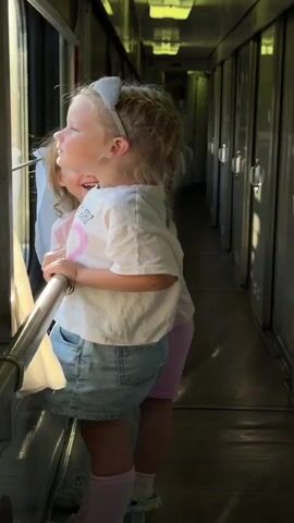 The video shows two young girls inside a train car. The girl on the left is wearing a pink t-shirt and pink shorts. She is laughing and holding onto a metal railing. The girl on the right is wearing a white t-shirt, denim shorts, and a headband with cat ears. She is also holding onto the railing and looking out the window. The train car is narrow with seats and windows along the side. The lighting suggests it is daytime. The girls are interacting with each other and the environment, appearing excited and engaged with their surroundings.