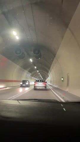 A vehicle is driving through a tunnel. The tunnel is lit with overhead lights. There are other vehicles in the tunnel, including cars ahead and to the left. The tunnel walls are smooth and curved, forming a cylindrical shape.