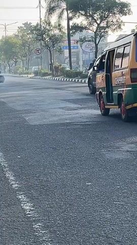 A yellow and green van drives down a paved road. The van has a red design with what appears to be a cartoon character on the rear window. Two people on motorcycles follow behind the van, one directly behind the van and another to the right of the van, slightly ahead. Trees line the road on the right side, and a few buildings are visible in the distance. The sky is clear and bright, suggesting daytime.