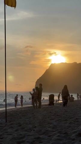 The video shows a sunset scene at a beach. The sun is setting behind a large hill or mountain, casting a golden glow over the scene. There are several people on the beach, most of whom are standing or walking. One woman is walking in the foreground, wearing a bikini and holding a phone. Other people are standing near the water, some appearing to take pictures. A red and yellow flag is planted in the sand in the foreground. The sky is a mix of orange, yellow, and grey clouds. The beach is sandy, and there are small waves in the ocean.