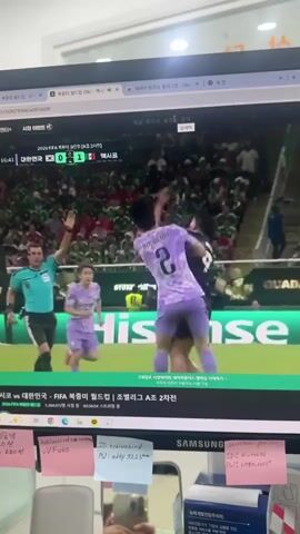 The video shows a computer monitor displaying a live soccer match. The score is South Korea 0, Mexico 1. A referee in a black and white striped shirt is on the field, along with two soccer players in purple and black uniforms. In the background, a crowd is visible. The video also shows a man's hand holding a smartphone, taking the picture. The monitor has sticky notes attached to its bezel. One note has the text "JD: aelly 8223."