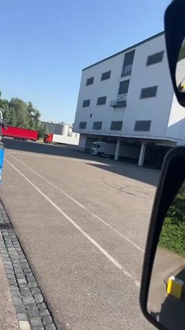 This media file captures a view from inside a truck cab, looking out at an industrial area in Mertingen, Germany. The scene is set on a bright, sunny day, with clear blue skies suggesting fair weather.

The primary subjects are trucks and a large, white industrial building with numerous square windows and ventilation units. The building appears to be a warehouse or factory. Several trucks are visible in the vicinity, including a red truck in the distance to the left and white semi-trailer trucks parked or maneuvering in front of the building. The truck cab itself is also visible, with side-view mirrors reflecting the surroundings, a steering wheel, and a smartphone mounted on the dashboard displaying a navigation app.

The activity taking place appears to be routine operations at an industrial site, possibly loading or unloading goods, or trucks arriving and departing. The time of day is indicated by the bright sunlight and shadows, likely midday.

Notable details include the clean, asphalted ground of the parking lot and road, marked with white lines. The side-view mirrors offer a slightly different perspective, showing greenery and a yellow object, possibly a storage bin, on the ground. The windshield of the truck shows some smudges, a common characteristic of vehicle glass. The smartphone screen displays "18:54" as the time, suggesting it's late afternoon. The navigation app shows a route with a destination labeled "Königssteiner Straße" and a distance of "7,39 km". There is also a small sign on the dashboard with what appears to be a company logo or name, possibly "TELEFUNKEN", though it is partially obscured.

Overall, the media conveys a functional and everyday scene within a logistics or manufacturing hub, captured from the perspective of a truck driver.