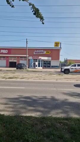 The video shows a street view with a car service building. The building has large garage doors and signs that say "АВТОСЕРВИС PRO" and "OIL." A white pickup truck with orange and gray graphics is parked in front of the building. A black car is parked in one of the garage bays. Traffic is visible on the street, including several cars and a trolleybus. The scene is set on a sunny day with trees and foliage in the foreground.