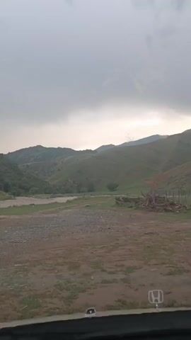 The video captures a scenic view of rolling green hills under a cloudy sky, likely from the perspective of a vehicle. The foreground is a dirt and gravel road, with some grass and debris. To the left, a dry riverbed or stream is visible. In the distance, the hills are covered with vegetation, with some bare patches and exposed earth. There are some logs and fencing visible on the right side of the road. The sky is overcast, with hints of light breaking through in the distance, suggesting either early morning, late evening, or an approaching storm. The camera appears to be moving, as indicated by the slight shift in perspective.