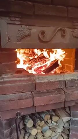 A video shows a brick fireplace with a fire burning inside. The fire is made up of several pieces of firewood that are engulfed in flames and glowing embers. Above the fire opening, there is a decorative metal plate with a grape vine design and a swirling pattern. Below the fire opening, there is a storage area filled with neatly stacked firewood. The camera is positioned in front of the fireplace, looking directly into the fire. The overall impression is one of warmth and coziness, suggesting a traditional hearth.