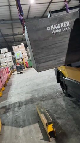 In an industrial setting, a yellow forklift, with a WLL (Working Load Limit) of 3,200 kg indicated on the yellow overhead bar, is lifting a large, grey, rectangular object branded "HAWKER Water Less" using purple straps and chains. The object is suspended in mid-air above the forklift. The background shows stacks of packaged goods, a green bin, and other industrial equipment. The floor is concrete and appears stained. The lighting is provided by overhead industrial lights. There are no people visible in the frame. The forklift seems to be in operation, presumably moving the heavy object.