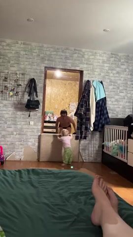 The video shows a room with a grey brick wall behind a green bed, where a young child is standing, a man appears to be getting dressed in the doorway, and another person is lying on the bed. A child, wearing green pajama pants and a pink t-shirt with a cartoon print, is the focus of the shot as they take a few steps and briefly hold a small object. In the doorway behind the child, a man with glasses and wearing dark underwear is visible. He appears to be taking off a shirt. The person holding the camera is lying on a green bed, visible only from the knees down, in the foreground. Behind the bed, along the wall, is a white crib with stuffed animals inside. A black bag and other items hang on the brick wall. The lighting in the room is bright.
