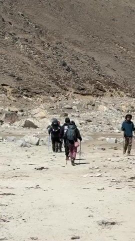 The video captures a group of trekkers hiking on a wide, dusty, and rocky trail amidst a stunning mountainous landscape, likely in Khumjung, Nepal. In the foreground, a person wearing a blue jacket and a large yellow and green backpack walks away from the camera, using trekking poles. Ahead of them, several other trekkers, also equipped with backpacks and trekking poles, proceed along the same path. The path is flanked by barren, rocky slopes, and in the distance, imposing snow-capped peaks rise against a bright blue sky with scattered white clouds. The sun is shining, casting clear shadows, indicating daytime with fair weather conditions. The scene depicts a high-altitude trek, with the individuals steadily making their way through the rugged terrain.