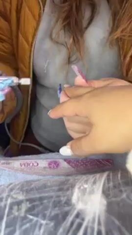 A woman is shown with a nail drill in her right hand. She has done nails with a shimmery finish, rhinestones, and blue, purple, and clear designs. The nail drill is pink. In her left hand, she is holding the hand of a woman whose nails have a white tip. The woman with white nails is wearing a yellow puffer jacket and a light gray top. She sits at a table covered in a fuzzy gray towel. Underneath the hand she is holding is a nail file, lying on the towel. The nail file is pink with the word "COBA" printed on it. The woman holding the nail drill works to prepare the nails for future manicures.