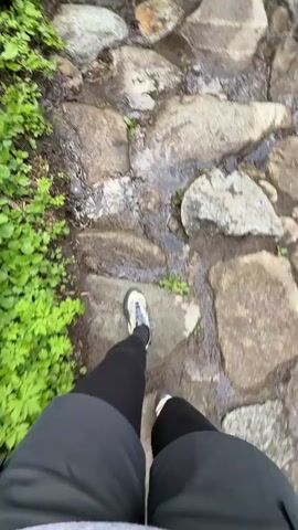 The video shows a first-person perspective of someone walking on a rocky path. The person is wearing black leggings and beige and grey hiking shoes. The path is made of various-sized grey rocks with small puddles of water in between. There is green foliage on the left side of the path.  The camera is focused downward at the feet and the path directly ahead.  The person appears to be moving forward at a moderate pace, navigating the uneven terrain.