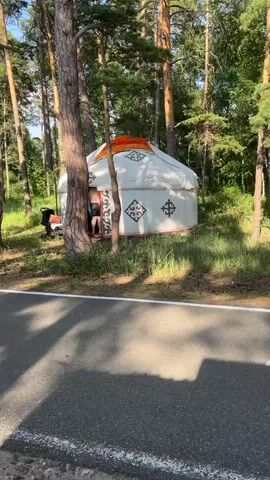 The video shows a yurt set up in a wooded area near a paved path. A person, dressed in dark shorts and a light-colored shirt, is partially visible entering the yurt. A stroller is positioned to the left of the yurt entrance. The yurt is white with orange accents on its roof and decorated with dark brown geometric patterns on its sides. Tall trees with rough bark surround the yurt, and grass and smaller plants grow around its base. The paved path in the foreground has a white line marking its edge. The lighting suggests it is daytime.