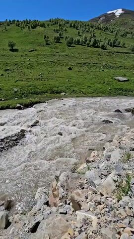 A flowing river runs through a rocky, grassy area, with a green mountain in the background. The sky is blue and clear. The mountain has some snow on its peak. There are trees scattered across the mountainside. The camera pans slowly to the right.