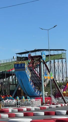 This video shows an amusement park ride with a pirate ship-themed gondola swinging back and forth. The ride is supported by a large metal structure, and there are other rides and structures visible in the background. The sky is clear and blue, suggesting it is a sunny day. The ride appears to be in operation, with people visible inside the gondola. The area around the ride is marked with red and white tire barriers, likely for a go-kart track or similar attraction.