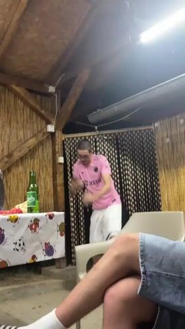 A young man wearing glasses and a pink t-shirt with white lettering and white shorts dances energetically in an outdoor covered area. He is standing near a table covered with a white cloth featuring colorful cow and fruit graphics. A green bottle and other items are on the table. Behind him, a dark curtain with a pattern hangs. In the foreground, a person's legs, wearing denim shorts and white socks with black stripes, are visible, facing away from the camera. The man in the pink shirt gestures with his hands while dancing.