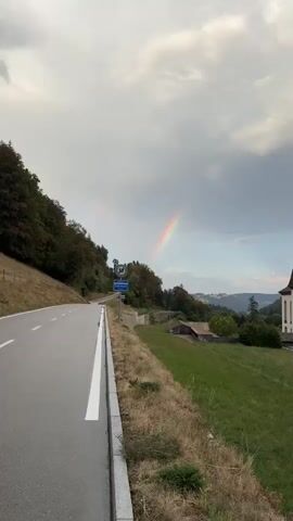 A vertical video pans right showing a road that curves to the left and away from the camera. On the left side of the road, a grassy embankment slopes upwards, covered in trees in autumnal colors. A white line marks the edge of the road. A speed limit sign showing 50 and a town name sign are visible on a post next to the road. To the right of the road, a grassy field slopes down and away. In the distance, there are rolling hills and some buildings. A rainbow is visible in the cloudy sky.