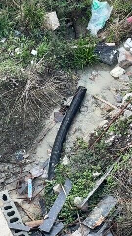 The video shows a black corrugated pipe lying in a shallow muddy puddle. The puddle is surrounded by grass, weeds, rocks, and trash, including a broken brick, a plastic bag, and a small black plastic crate. Water is flowing from the open end of the pipe into the puddle.