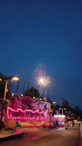 A video shows a carnival ride at dusk. The ride is brightly lit with pink and blue lights. Fireworks are exploding in the dark blue sky above the ride. Several people are visible walking on the path in front of the ride. The ride appears to be a carousel with decorative elements. Streetlights illuminate the area.