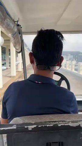 The video shows a person driving a golf cart on a pier. The driver is wearing a navy blue polo shirt and glasses. The golf cart is moving along the pier, and the ocean and a lighthouse can be seen in the background. The weather appears to be overcast.