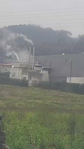 The video shows an industrial complex emitting smoke. The complex consists of large, low-rise buildings, some with visible chimneys. Smoke is rising from the complex, indicating industrial activity or potentially a fire. A grassy field lies in the foreground, with a blurry, possibly forested, hillside visible in the background under a hazy sky. Power lines run across the top of the frame.