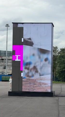 This video shows a large, rectangular LED screen displaying abstract art. The screen is positioned outdoors in an asphalt area, with a person standing to the left of it. In the background, there is a tall utility pole, a building under construction, and some trees. The screen displays various images and colors that change over time, including what appears to be a close-up of fabric, a red line, and red circular shapes falling from the top. The screen also has a section on the left with geometric shapes in pink and white.