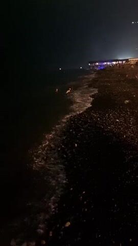 A nighttime view of a pebble beach. The dark water of the sea laps at the shore, with gentle waves breaking. Several people are visible in the water, some swimming or wading, while others are sitting or lying down near the water's edge. On the beach, a person is standing near the water, and further up the beach, lights indicate a building or a strip of businesses. The overall scene is dark, illuminated only by distant artificial lights and the faint glow of the water.