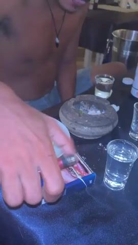 This media file captures a person, seemingly male, sitting at a table in what appears to be a restaurant or bar setting. The individual is shirtless, wearing denim shorts, and has blonde-dyed hair. They are wearing a necklace with a dark pendant.

On the table, there are several shot glasses filled with a clear liquid, a small mortar and pestle containing ash and cigarette butts, a pack of cigarettes, and an empty can. The table is covered with a dark, textured cloth. In the background, other people can be seen at tables, and there are overhead lights with woven shades, suggesting an indoor or covered outdoor evening setting. The general ambiance suggests a casual, social environment, possibly a tourist destination given the context of Preah Sihanouk, Cambodia.

The individual's facial expression and posture, with their tongue slightly sticking out, could imply a moment of relaxed enjoyment or a playful pose. The presence of alcohol and cigarettes points to a social gathering or a personal moment of unwinding. The lighting is warm, characteristic of evening illumination in a commercial establishment. No visible text is present in the media.