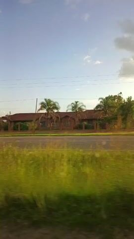 The video shows a view from inside a moving vehicle, looking out a side window. The scenery consists of a road and some vegetation along the side of the road. There are also buildings and billboards visible in the background. A few cars and a motorcycle pass by on the road. The sky is blue with some white clouds. The general setting appears to be a rural or suburban area, possibly near a factory or industrial complex based on the billboard text that reads "Nspra Factory." There are no people clearly visible within the view of the window of the vehicle.