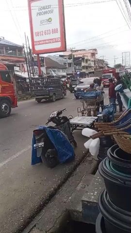 The video shows a street scene with vehicles and people. A large red truck with "PERTAMINA" written on it is visible, along with other cars and motorcycles. There are several people standing on the sidewalk and near the vehicles. Objects such as plastic containers, baskets, and gardening tools are displayed on the sidewalk. A motorcycle covered with a blue tarp is parked on the side of the road. A billboard advertising "LOKASI PROMOSI" is also visible. The overall impression is of a busy urban environment with commercial activity.