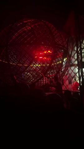 The video shows a "Globe of Death" motorcycle stunt at a show. A large, spherical cage dominates the scene, lit by red and white lights as two or more motorcyclists zoom around inside, visible as glowing lights within the structure. The audience watches from below, their silhouettes barely visible in the dark foreground. A person dressed in red walks alongside the cage, and the backdrop features painted scenery.