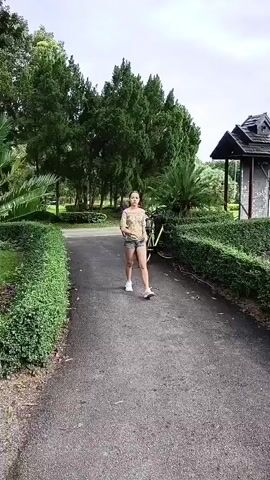 A young woman in a patterned shirt and denim shorts walks towards the camera on a paved path. A small child wearing a floral dress walks towards the woman and reaches out to touch her. The background features manicured bushes, trees, and a gazebo-like structure.