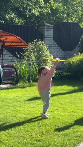 A young girl with pigtails is standing on a grassy lawn, launching a yellow toy airplane. She is wearing a light pink sweatshirt, grey pants, and a headband. The airplane takes off and then quickly descends to the ground. A person's arm is briefly visible to the left of the frame. A dark-colored wooden fence with brick pillars runs along the background. Various bushes and trees are scattered throughout the yard, and an orange canopy is visible in the left corner. The scene is brightly lit by sunlight, casting shadows across the grass.