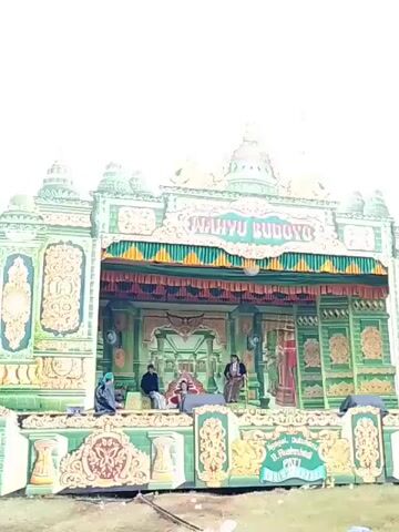 Here is a description of the video:

The video shows a performance stage in an outdoor setting.  The stage is decorated in green and gold, with the words "WAHYU BUDOYO" written across the top.  Four people are on the stage. One person is seated on what appears to be a throne, another person is seated to the side of the "throne." Two people are sitting on the edges of the stage. The video is filmed from a distance and is slightly overexposed.