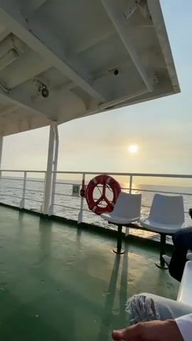 The camera pans slowly over a section of a ferry deck. The deck is green and covered by a white overhead structure. The sun is low in the sky, shining over the ocean. A red life preserver is attached to the railing next to two white plastic chairs. Two people are seated in the chairs. Only their lower legs and part of their arms are visible. One person wears ripped jeans, the other wears slacks and a white dress shirt.