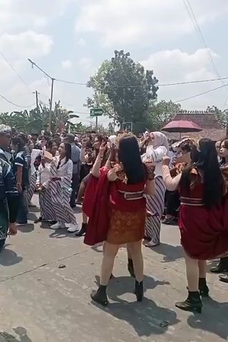 The video shows an outdoor street scene with a crowd of people watching a performance by several women dressed in traditional attire. The performers are wearing short, ornate dresses in red and gold patterns, with accompanying capes and head coverings. They appear to be dancing or performing a choreographed routine. Some of the onlookers are also dressed in traditional striped fabrics, suggesting a cultural event or festival. The environment is a paved street with buildings and trees visible in the background, under a bright, sunny sky. There are power lines overhead. The performers are the primary focus, with the crowd forming a backdrop to their activity. The mood appears festive and lively.