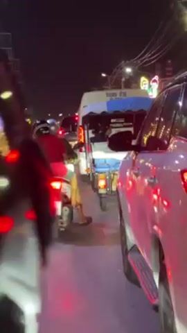 At night, a congested street scene is depicted, with traffic moving slowly.  The camera perspective is from within a stationary vehicle, likely a car, on the right side of the road. In front, a white auto rickshaw ("tuk-tuk") carries passengers. A man wearing a red shirt and riding a scooter is also in front of the camera vehicle. Many motorcycles and a white pickup truck also populate the road. Ambient lighting and illuminated signs suggest a commercial area. Pedestrians can be seen along the sidewalks.