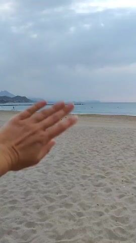 The video begins with a close-up of a person's hand waving. The hand is in the foreground and obscures a large portion of the frame. The background shows a sandy beach with the ocean and a cloudy sky. As the video progresses, the hand moves out of frame, revealing more of the beach and the ocean. A few people can be seen in the distance on the beach and in the water. The sky is overcast with scattered clouds. The overall scene is a tranquil beach setting.