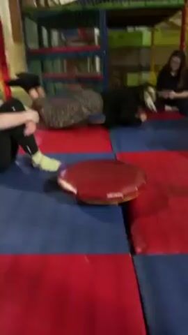 In a play area with a red and blue padded floor, three women are visible. One woman is sitting with her legs crossed and a phone in her hands. Another woman in black leggings and socks sits with her legs bent and arms around them. A third woman is on the floor, attempting to climb onto a red, circular platform with a yellow border. She first puts her hands and knees on the platform, then slips and crawls forward. Eventually, she successfully gets on the platform and then falls backwards, landing on her back with her legs in the air. The woman on the floor laughs as she watches the woman struggling with the platform. A play structure with multiple levels is visible in the background.