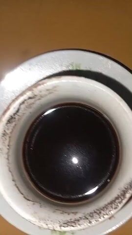 A close-up shot of a white teacup filled with dark liquid, possibly coffee or tea, resting on a matching saucer. The teacup has a slightly worn rim with some brown staining. The saucer has a delicate floral pattern. The lighting highlights the glossy surface of the liquid.