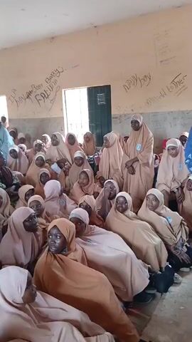 The video shows a group of young girls, likely students, sitting on the floor in a room. They are all wearing similar beige hijabs. There are also a few adult women present. One woman, wearing a blue patterned hijab and dress, is standing and appears to be addressing the girls. Another woman, wearing glasses and a white top over a green garment, is also standing and seems to be interacting with the woman in blue. The room appears to be a classroom, with a window and some writing on the walls. The lighting is natural, coming from the window. The atmosphere appears to be calm and orderly, with the girls attentively listening.