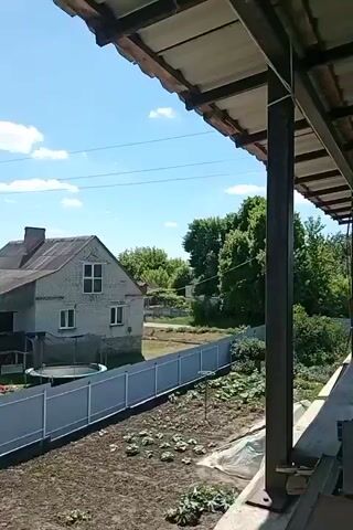 The video captures a panoramic view of a rural landscape on a sunny day with a blue sky and scattered white clouds. In the foreground, a gray fence runs horizontally, with a garden bed and some plants visible beyond it. Behind the fence, a two-story house with a gray roof and white walls stands prominently, with a trampoline in the yard. A power pole with overhead wires is situated near the house. The background reveals lush green fields and trees, with several other houses and structures visible in the distance, suggesting a small village or settlement. The camera pans slowly from right to left, offering a broader perspective of the surroundings.