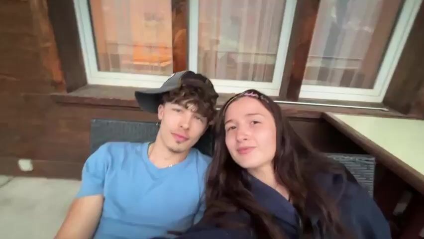 This media file features two individuals, a young man and a young woman, posing for a selfie on what appears to be a balcony or patio. The man, wearing a blue t-shirt and a baseball cap, sits on a dark woven chair. The woman, with long dark hair and a headband, is seated next to him, leaning back with her eyes closed. The background shows the exterior of a wooden building with windows and curtains.

The scene then shifts to a wider view of a mountainous landscape, likely Grindelwald, Switzerland. The perspective focuses on a person's legs and feet, wearing grey socks, resting on a balcony railing. Below, a picturesque village is nestled amongst green hills, with majestic, snow-capped mountains dominating the background. The weather appears overcast with a cloudy sky. The overall mood conveyed is relaxed and serene, possibly indicating a vacation or a peaceful moment enjoying the scenery.