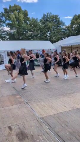 A group of young women in black dresses and white sneakers are performing a synchronized dance routine on a wooden outdoor stage. They are arranged in two rows, facing the camera. The woman in the front row on the left is wearing a blue bow in her hair. The dancers are kicking their legs and moving their arms in unison. In the background, there are white tents and trees. The sky is blue with some clouds.