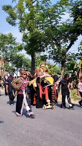 A procession is moving down a paved road. In the center of the frame, a large, elephant-shaped costume is being pushed and pulled by several people. The elephant costume is black with yellow and red accents. A person in elaborate traditional clothing, possibly a headdress and crown, is seated on top of the elephant costume. Around the elephant, other individuals are walking, dressed in black clothing with red accents. Some are holding long poles. The environment appears to be outdoors with trees lining the road and a clear sky. The overall atmosphere suggests a cultural event or parade.