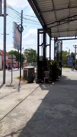 A video shows a street scene from a sidewalk, with a focus on utility poles and overhead wires. In the background, there are buildings, including a restaurant with a yellow sign that reads "WARUNG MAKAN MBAH MEN WIWIK," and several motorcycles are parked and moving on the road. The sky is visible with some clouds. The perspective is from ground level, looking across the street. The camera pans slightly throughout the video. There are no distinct people or interactions clearly captured.
