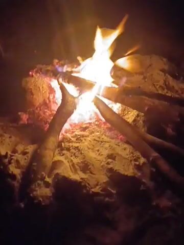 A campfire burns brightly in a sandy area. The fire is built in a teepee shape with three logs leaning into the center. Flames lick upwards, illuminating the sand around the base of the fire. The scene is dark, suggesting it is nighttime.