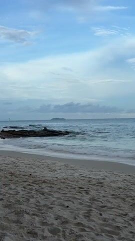 A tranquil beach scene unfolds under a vast, pale blue sky dotted with wispy clouds. The foreground is dominated by a sandy beach, its surface textured with subtle ripples. The gentle surf of the ocean laps at the shore, creating a thin line of foam that traces the coastline. A series of dark, rocky outcrops emerge from the water, some appearing wet and glistening from the waves. In the distance, the horizon is marked by the faint outline of a small island. A white boat, its sleek form cutting through the water, is visible on the left side of the frame, moving away from the viewer. On the far left, the dark silhouette of a tree, with its branches reaching out, frames the scene, partially obscuring a wooden object in the bottom left corner. The overall impression is one of peaceful serenity, with the soft light suggesting either early morning or late afternoon.